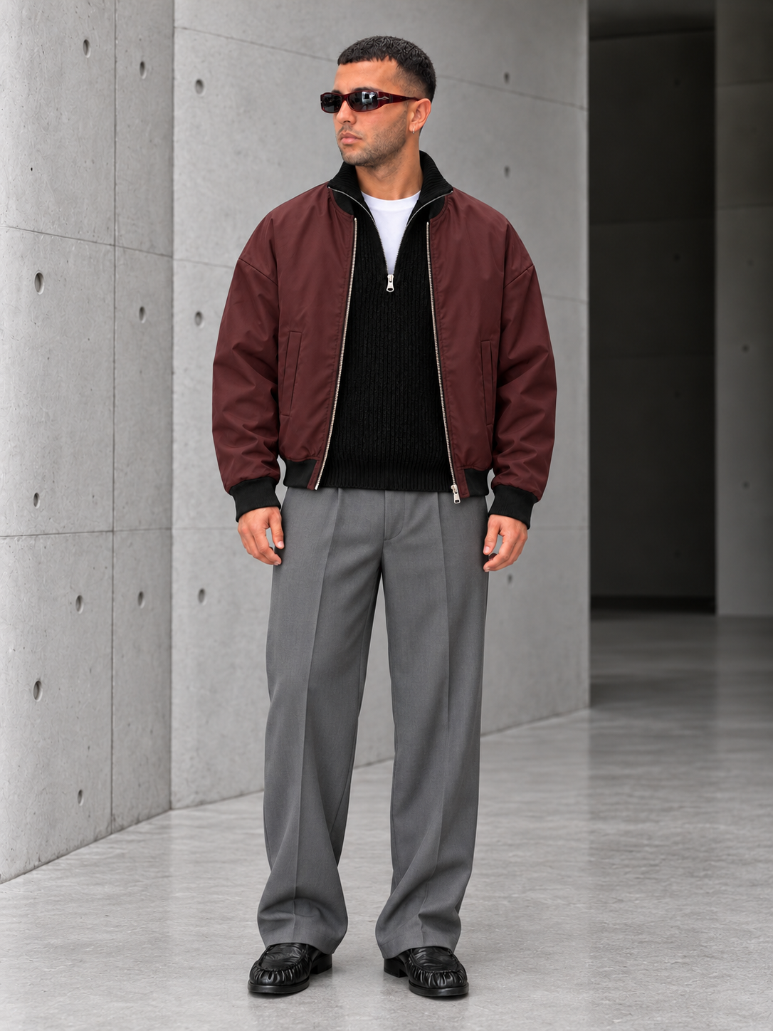 Outfit con bomber bordeaux, pullover nero e pantalone grigio sul cemento